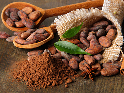 a number of raw cocoa beans in a sack and spoons alongside cocoa powder