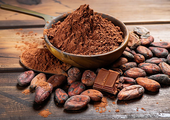 a spoon of cocoa powder sat on top of cocoa beans an two pieces of chocolate